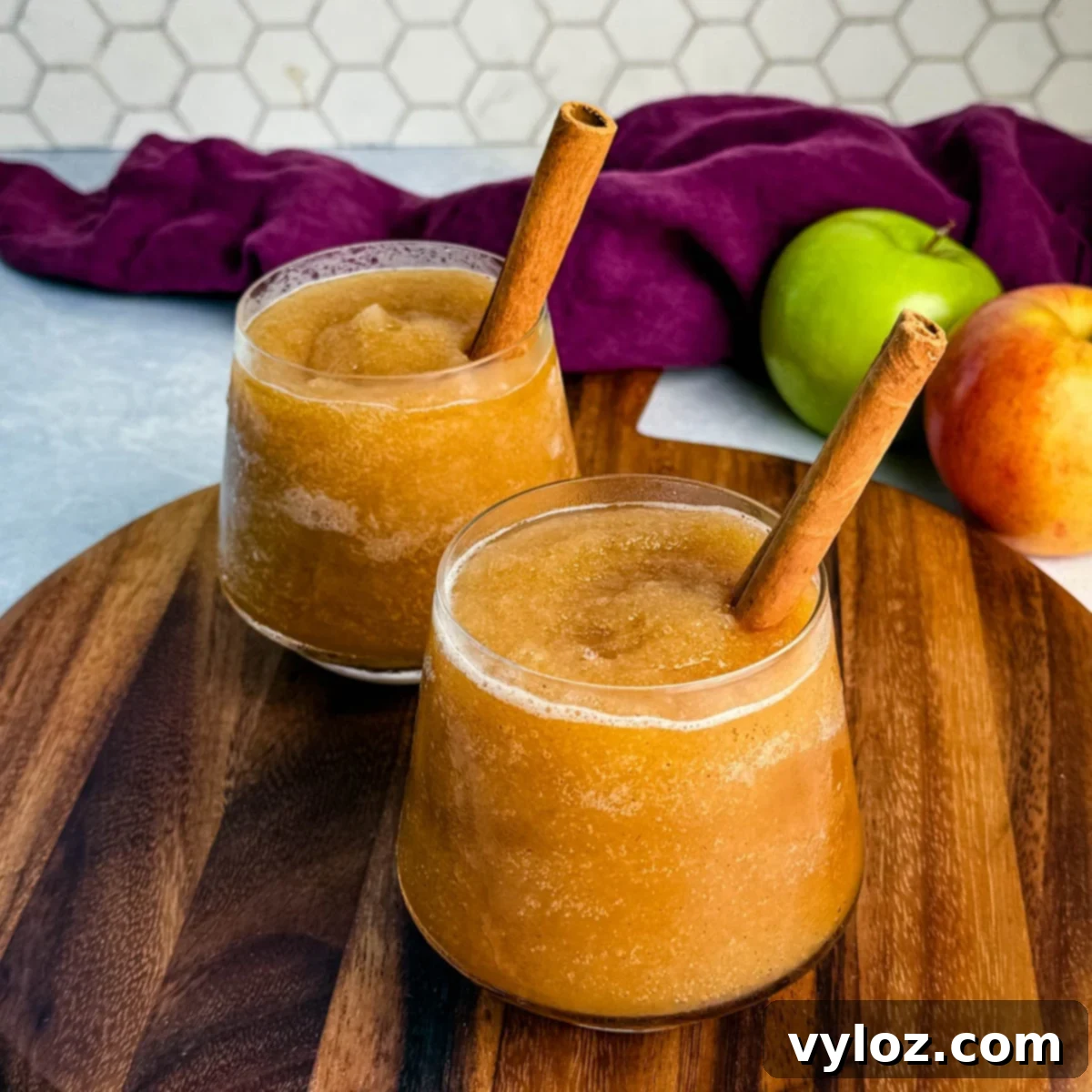 Two glasses of frozen apple cider slush served on a wooden board, each garnished with a cinnamon stick, with apples and a purple cloth in the background.