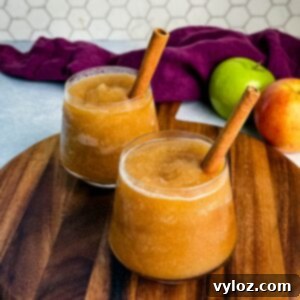 Two glasses of frozen apple cider slush served on a wooden board, each garnished with a cinnamon stick, with apples and a purple cloth in the background.