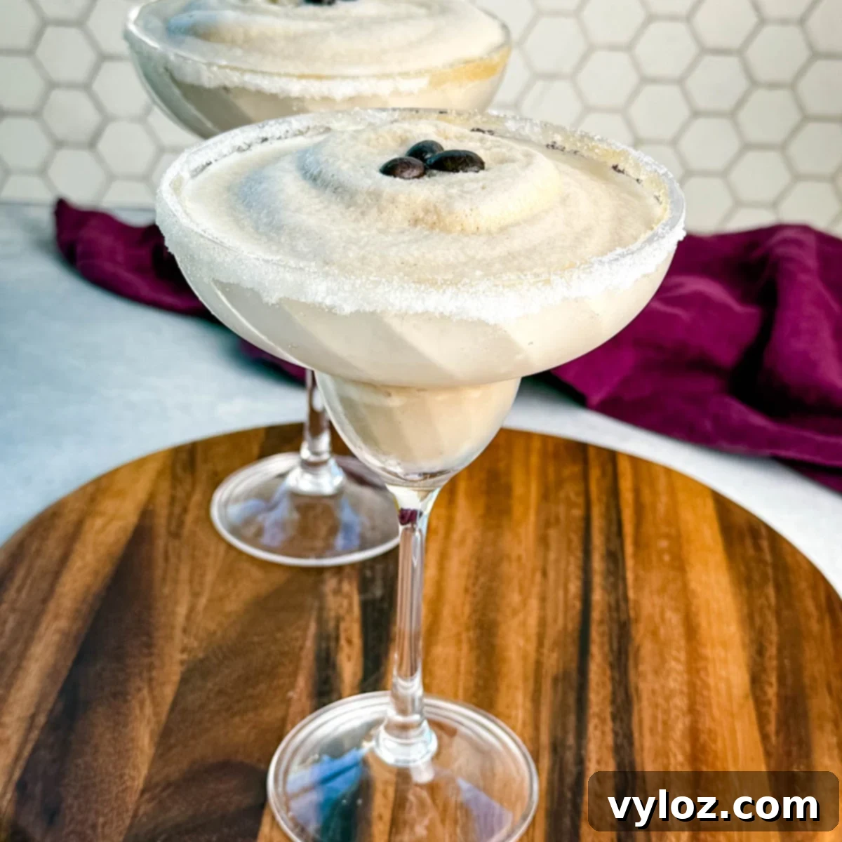 Frozen Shadow Espresso Martini 2 Two frozen espresso martinis in sugar-rimmed glasses topped with coffee beans, served on a wooden board with a purple cloth in the background.