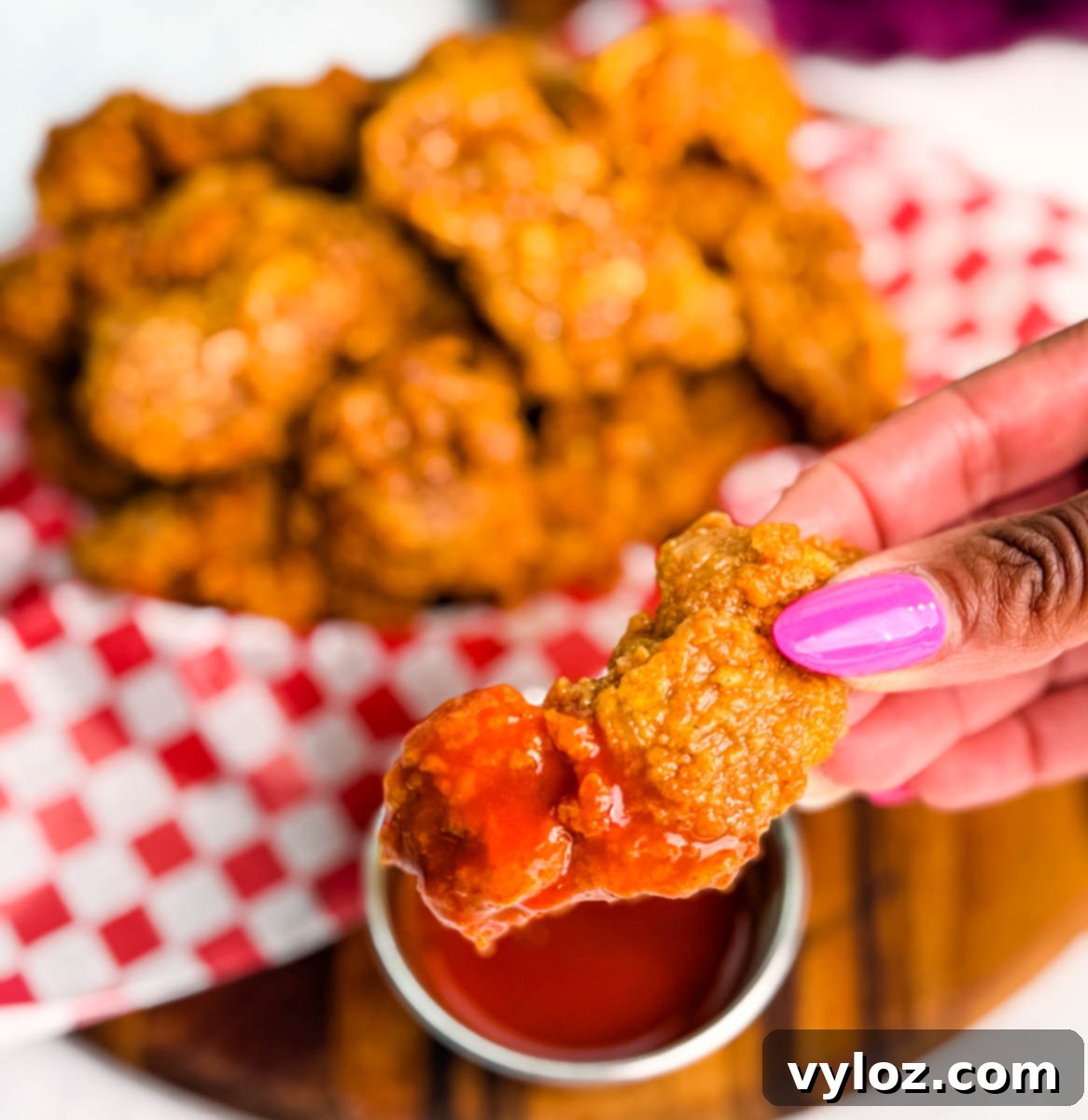 Close-up of a hand holding a crispy fried chicken gizzard dipped in bright red hot sauce, with a basket of fried gizzards blurred in the background.