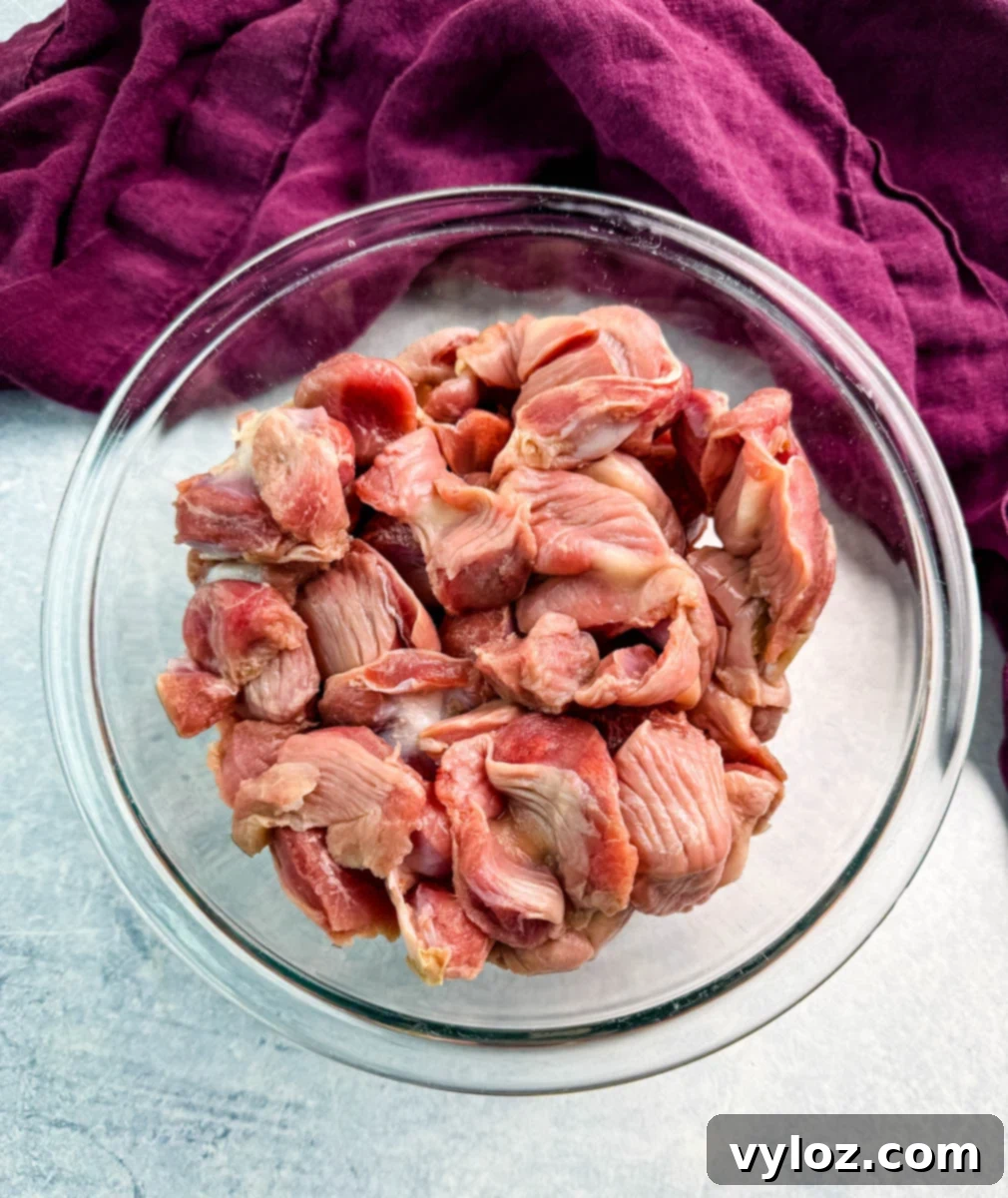 Overhead view of raw chicken gizzards in a clear glass bowl, set on a light background with a purple cloth nearby.