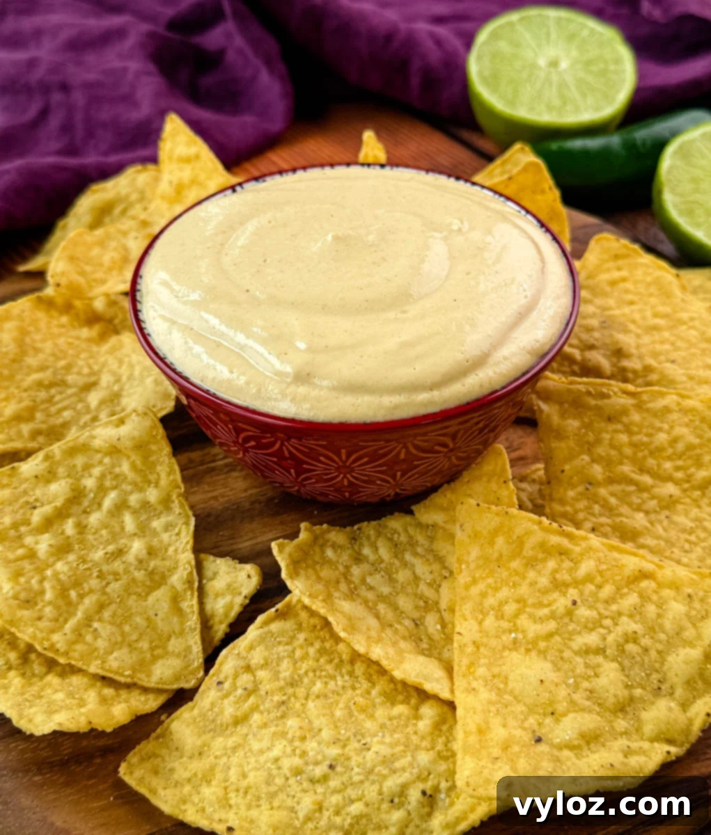 A decorative bowl filled with creamy high-protein queso dip on a wooden board, surrounded by crispy tortilla chips and garnished with lime and jalapeño.
