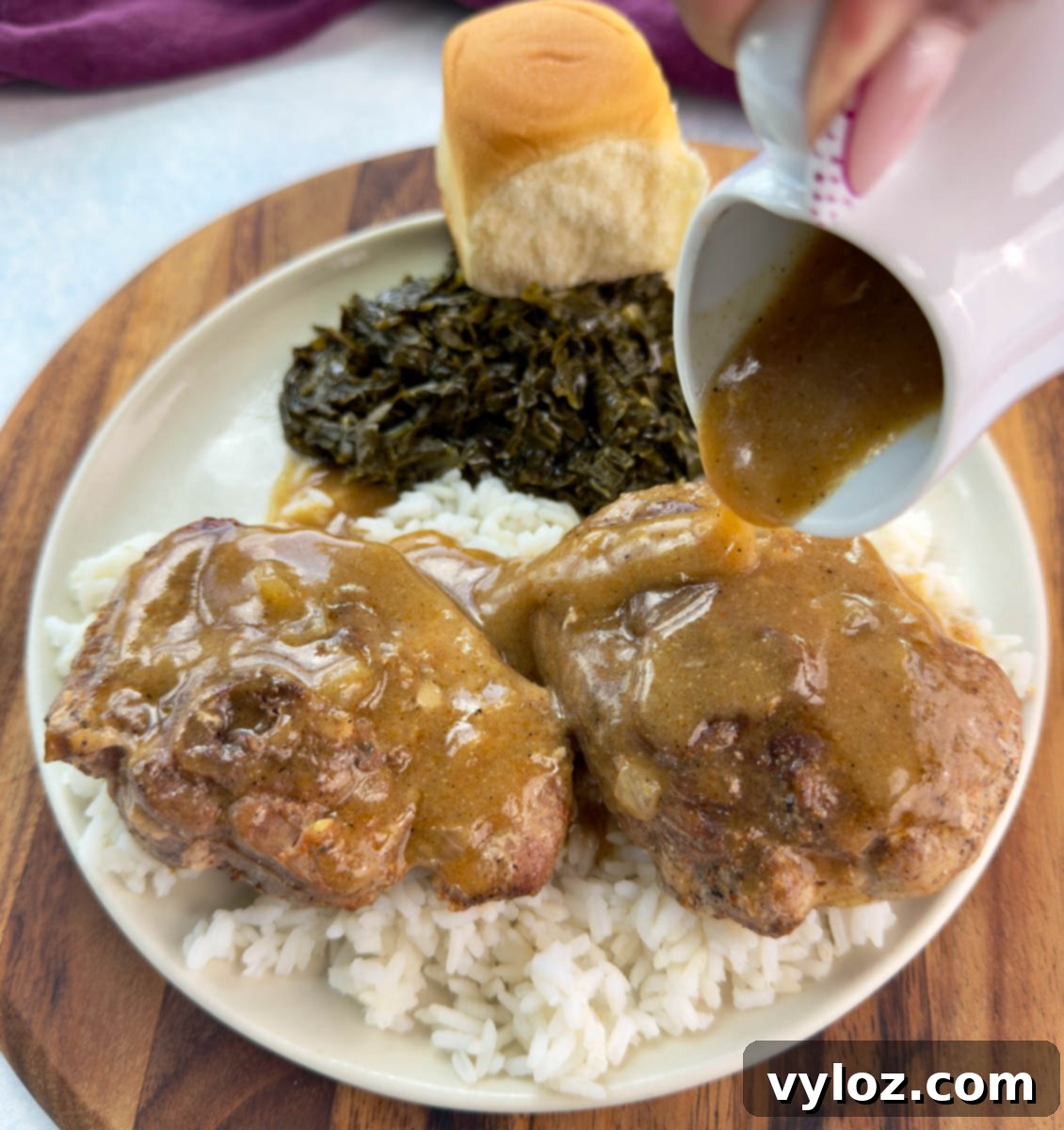 Close-up of gravy being poured from a white pitcher over turkey chops served with rice, collard greens, and a roll.