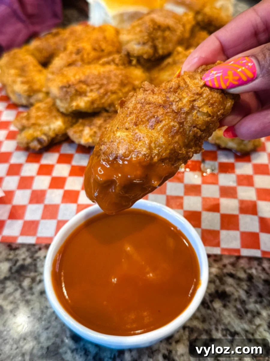 Hand holding a crispy fried chicken wing dipped into a bowl of bright orange buffalo sauce, with more fried wings in the background.
