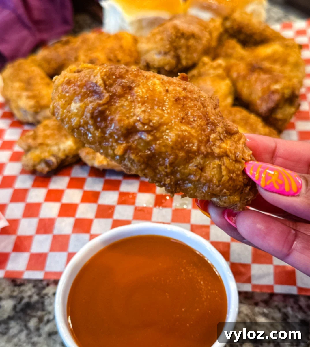 Hand holding a golden, crispy fried chicken wing over a small bowl of bright orange buffalo sauce, with more fried chicken pieces blurred in the background.