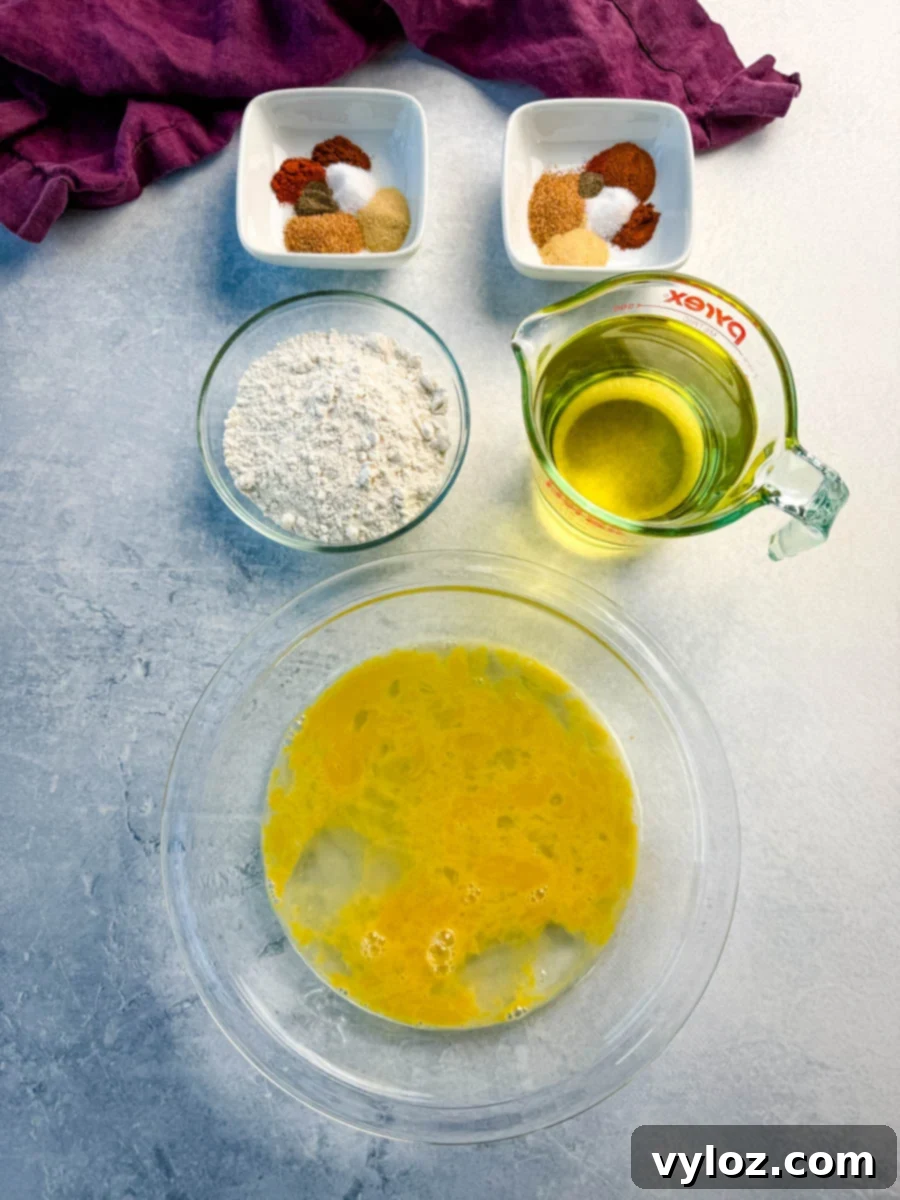 Overhead shot of fried chicken prep ingredients: a bowl of whisked eggs, a measuring cup of oil, a bowl of flour, and two small dishes of spice blends.