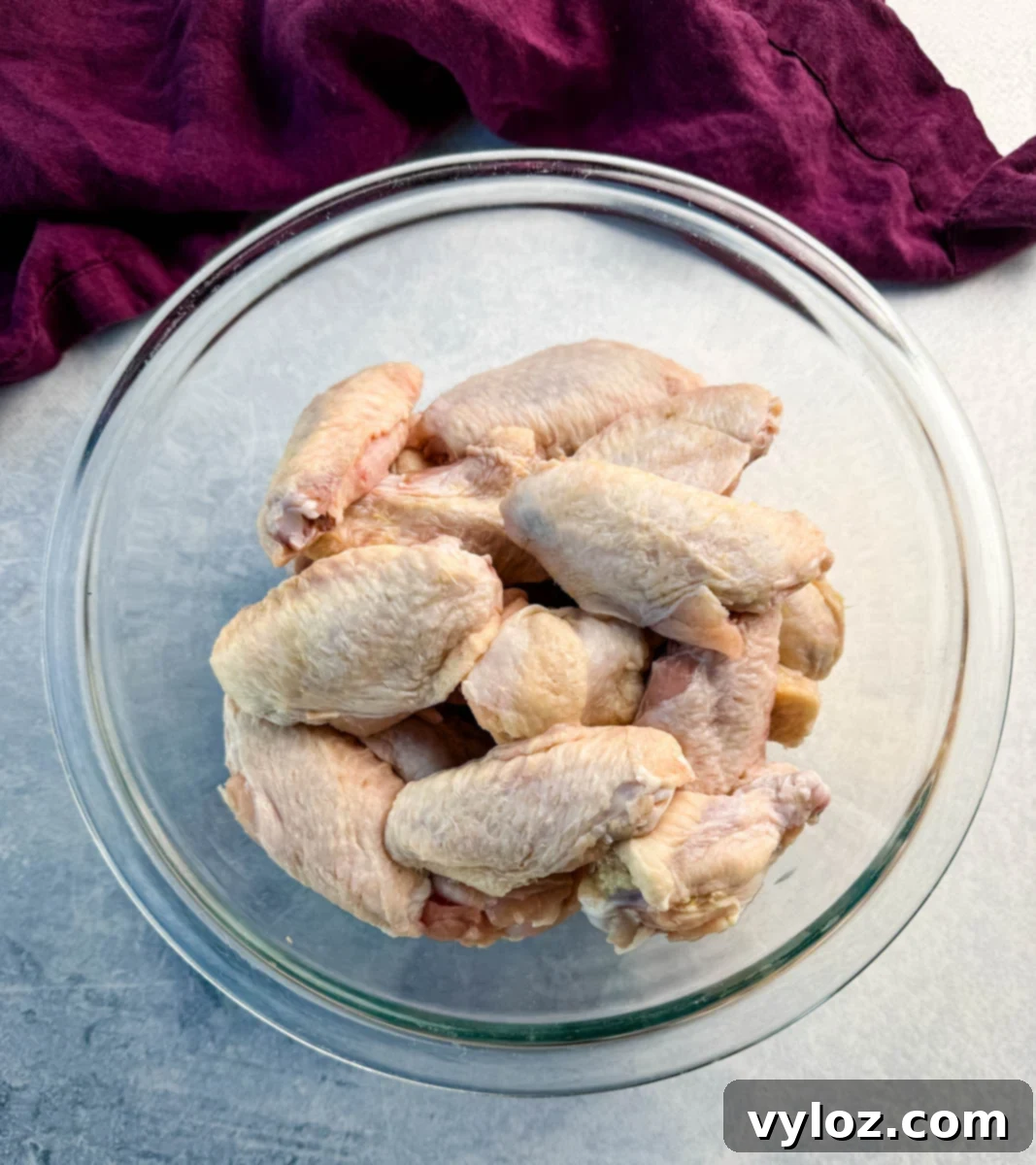 A clear glass bowl filled with raw chicken wings, placed on a counter with a purple cloth in the background.