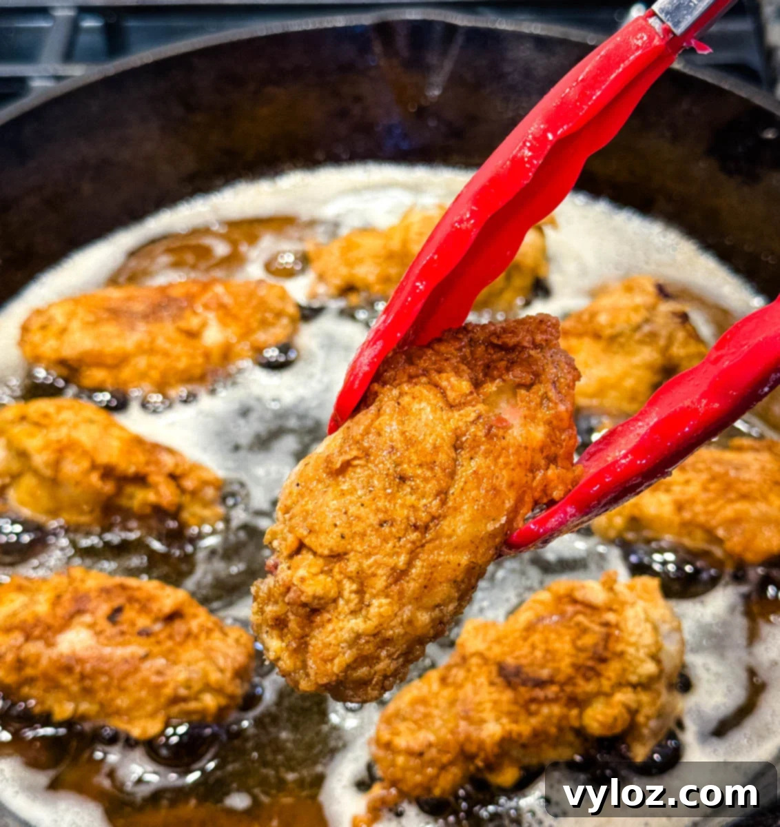 Crispy fried chicken wing held with red tongs over a skillet of bubbling hot oil with several pieces frying.