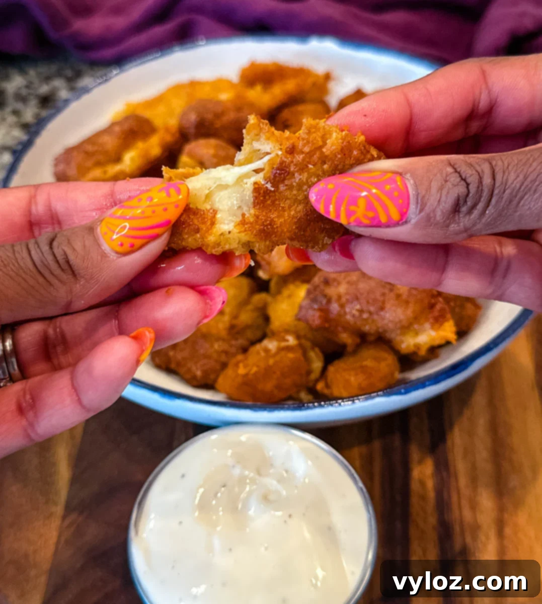 Close-up of hands pulling apart a crispy fried cheese curd to reveal gooey melted cheese inside, with a bowl of ranch dipping sauce below.