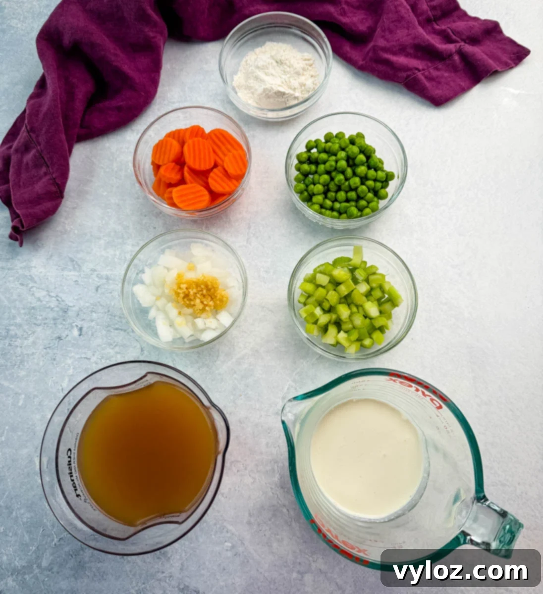 Bowls of prepped ingredients for chicken pot pie casserole — carrots, peas, celery, onion with garlic, flour, chicken broth, and cream — arranged on a gray surface with a deep purple kitchen towel in the background.