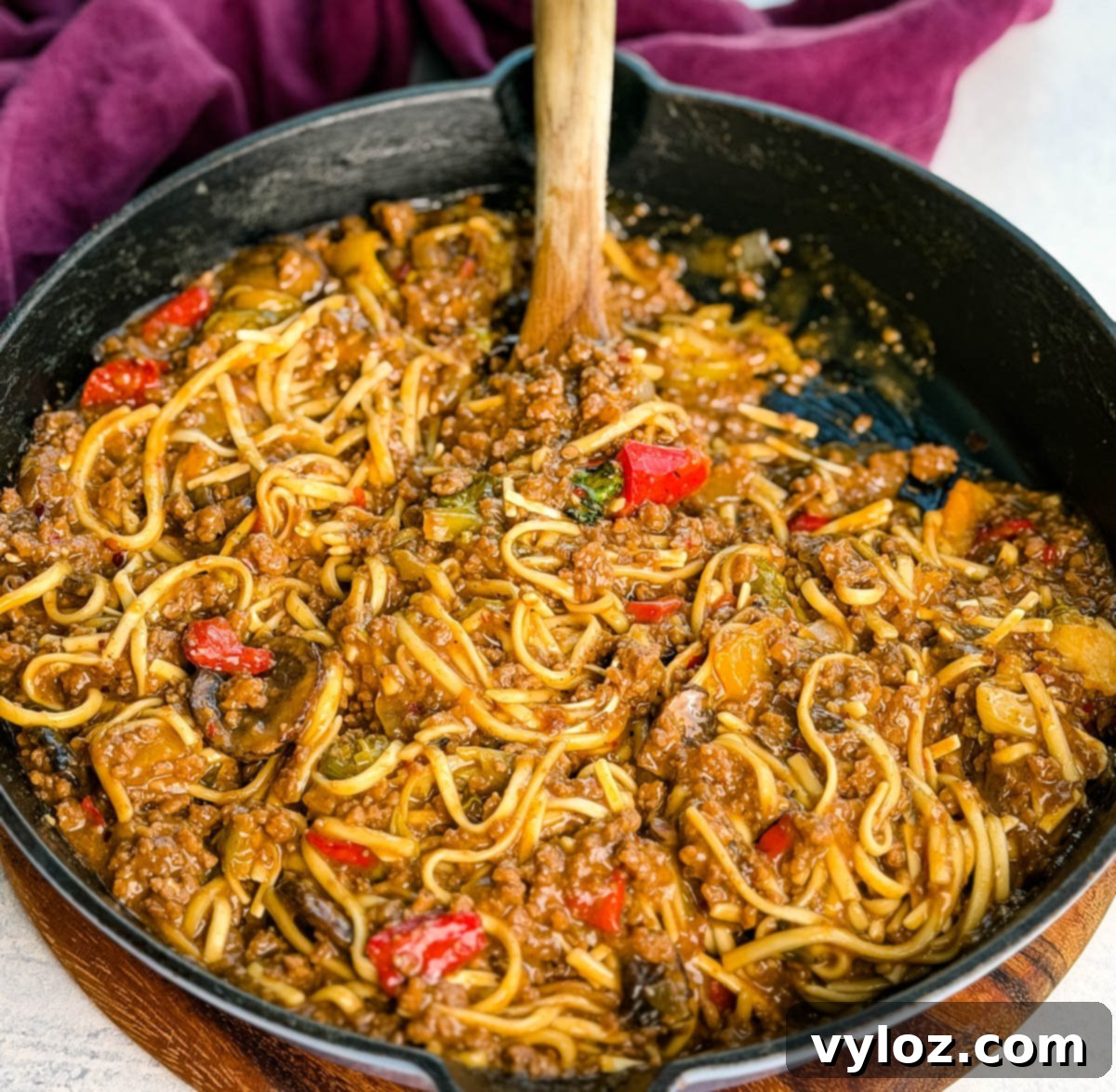 Overhead photo of the entire skillet filled with glossy Mongolian ground beef noodles, with a wooden spoon in the pan. A purple napkin sits in the background. The vibrant sauce and colorful veggies stand out against the black skillet, highlighting the dish's irresistible appeal.