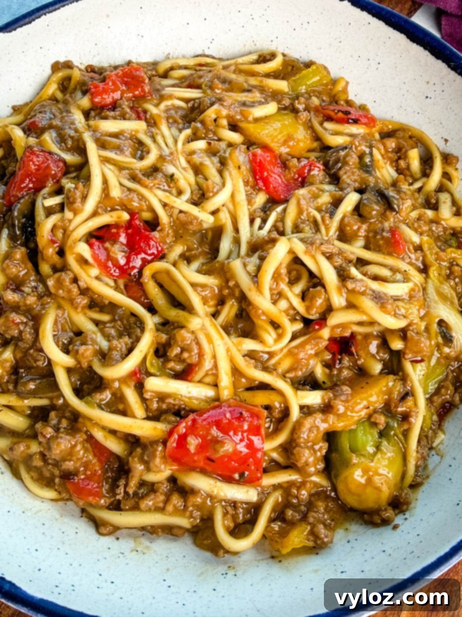 Close-up shot of Mongolian ground beef with udon noodles served in a white bowl with a blue rim. The noodles are coated in a rich brown sauce with red and yellow bell peppers mixed in, ready to be enjoyed.