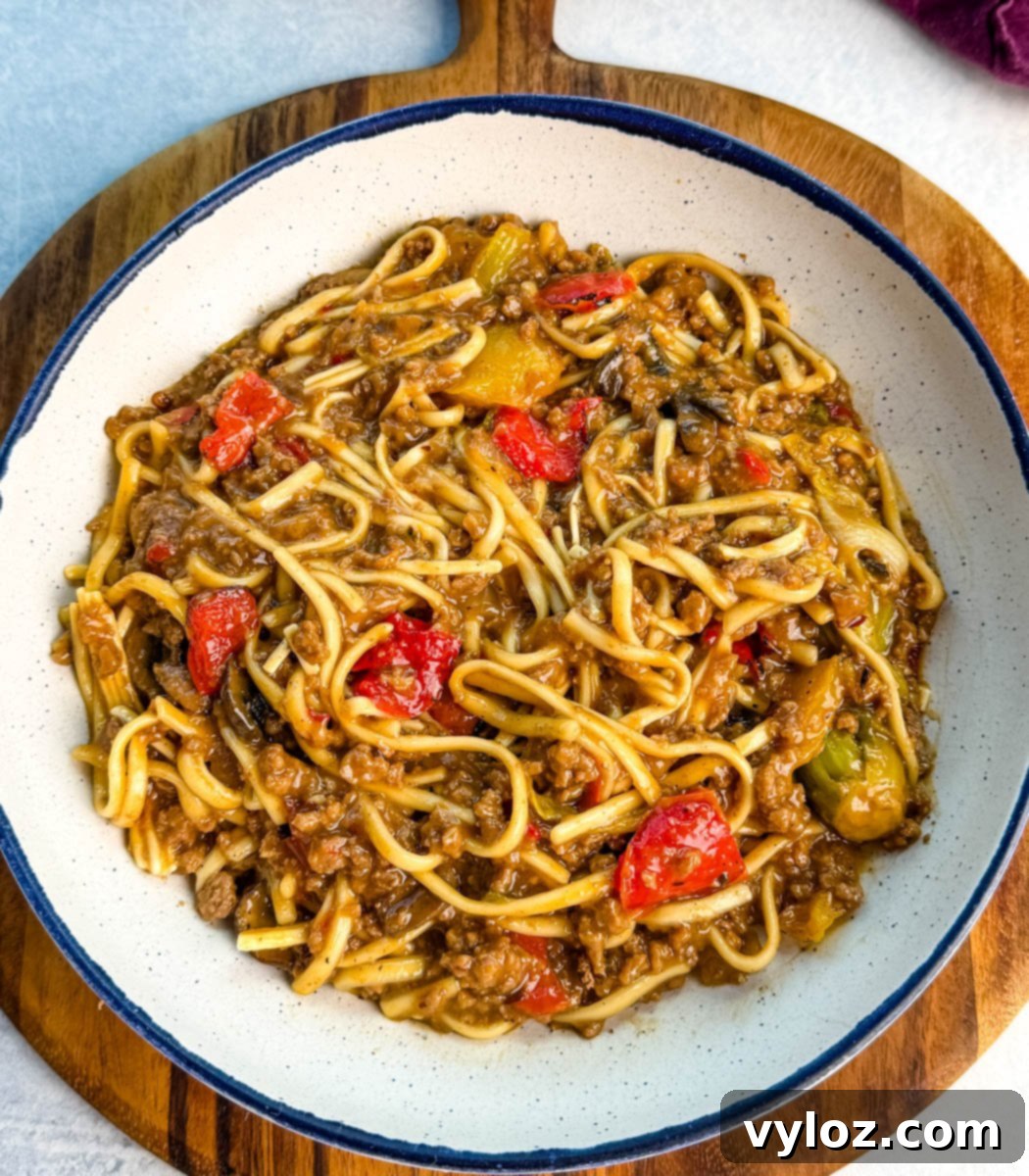 Overhead view of Mongolian ground beef and noodles in a white bowl on a wooden board. The dish looks saucy and hearty, with colorful vegetables and thick udon noodles, garnished for presentation.