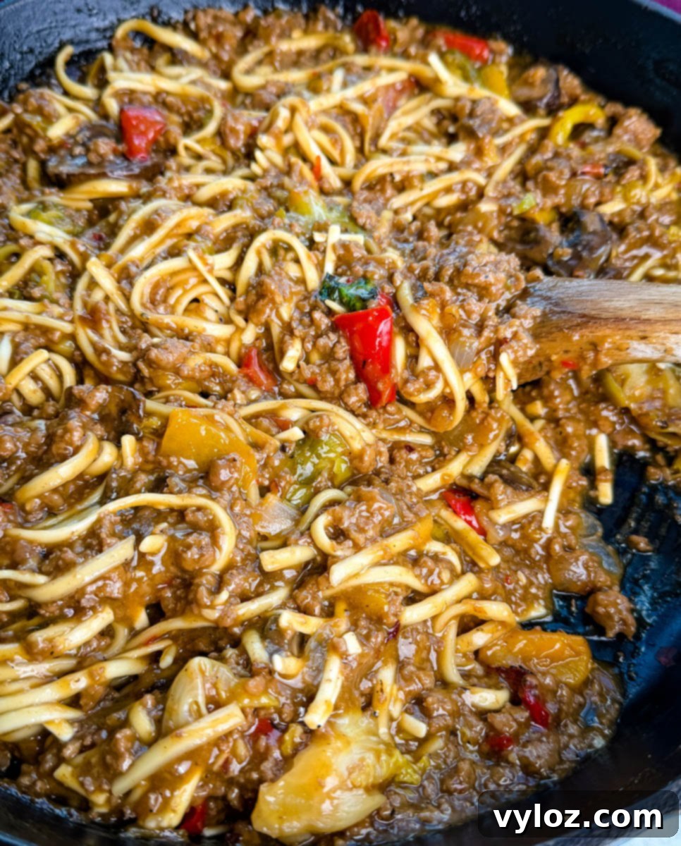 Close-up shot showing saucy Mongolian ground beef noodles with red peppers, mushrooms, and bits of green vegetables, highlighting the glossy, caramelized texture of the sauce. The steam rising from the hot dish adds to its inviting appeal.