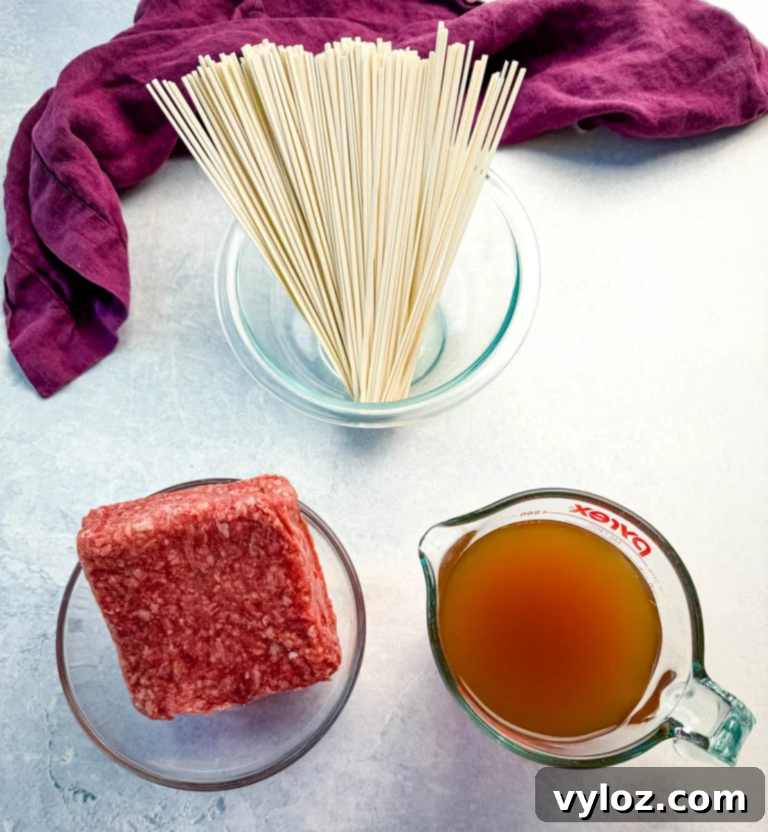 Overhead shot of three main ingredients for one pot Mongolian ground beef with udon noodles—raw ground beef, uncooked udon noodles in a glass bowl, and a measuring cup of broth. A purple kitchen towel sits in the background on a light surface, ready for cooking.