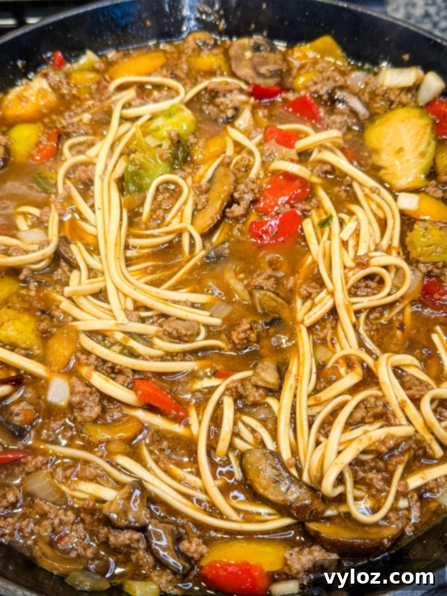 Close-up shot showing saucy Mongolian ground beef noodles with red peppers, mushrooms, and bits of green vegetables, highlighting the glossy, caramelized texture of the sauce and its rich color.