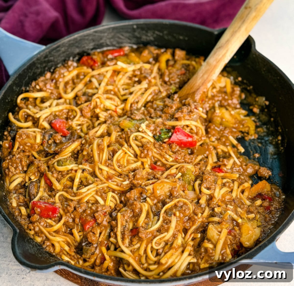 Overhead photo of the entire skillet filled with Mongolian ground beef noodles, a wooden spoon resting in the pan, and a purple napkin in the background. The glossy sauce and colorful veggies stand out vividly against the dark black skillet, inviting you to dig in.