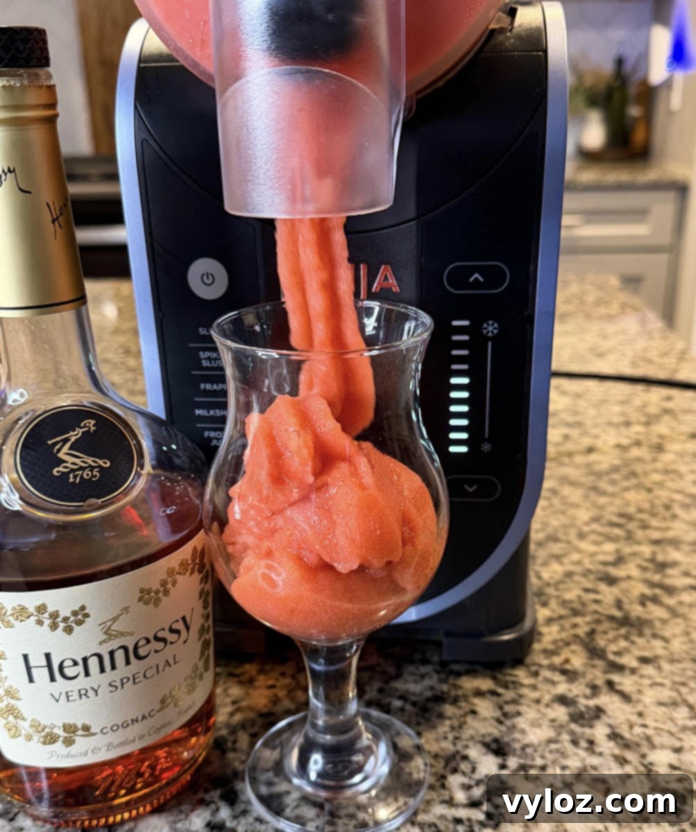 Hennessy Midnight Slush 8 Ninja Slushi machine dispensing a smooth red Hennessy slushie into a glass beside a bottle of Hennessy on a kitchen counter.