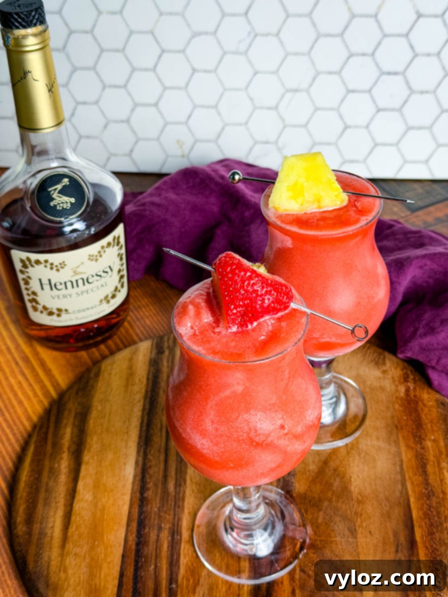 Hennessy Midnight Slush 9 Two bright red Hennessy slushies in curved hurricane glasses topped with a strawberry and pineapple slice, sitting on a wooden board with a bottle of Hennessy and a purple napkin in the background.
