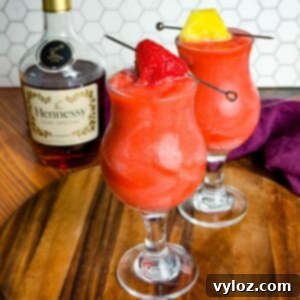 Hennessy Midnight Slush 10 Two bright red Hennessy slushies in curved hurricane glasses topped with a strawberry and pineapple slice, sitting on a wooden board with a bottle of Hennessy and a purple napkin in the background.
