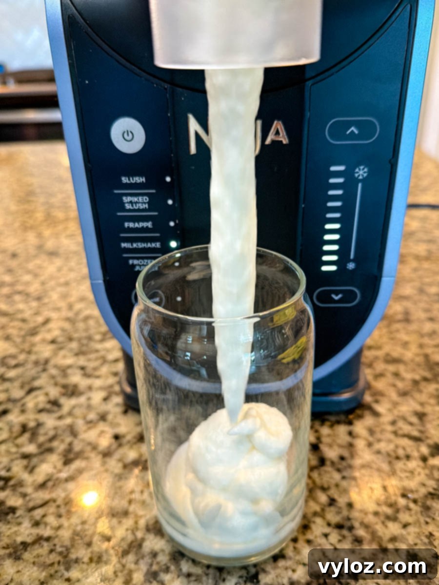Stealthy Slushy Delight 6 A creamy vanilla mixture dispensing from a Ninja Slushi machine into a clear glass on a granite countertop, showing the thick McFlurry-style texture forming as it pours.