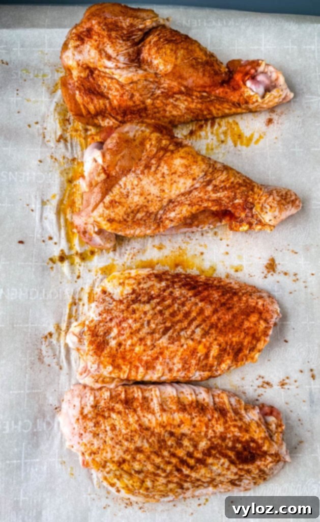 Raw turkey wings generously coated with seasoning and arranged on parchment paper, prior to cooking.