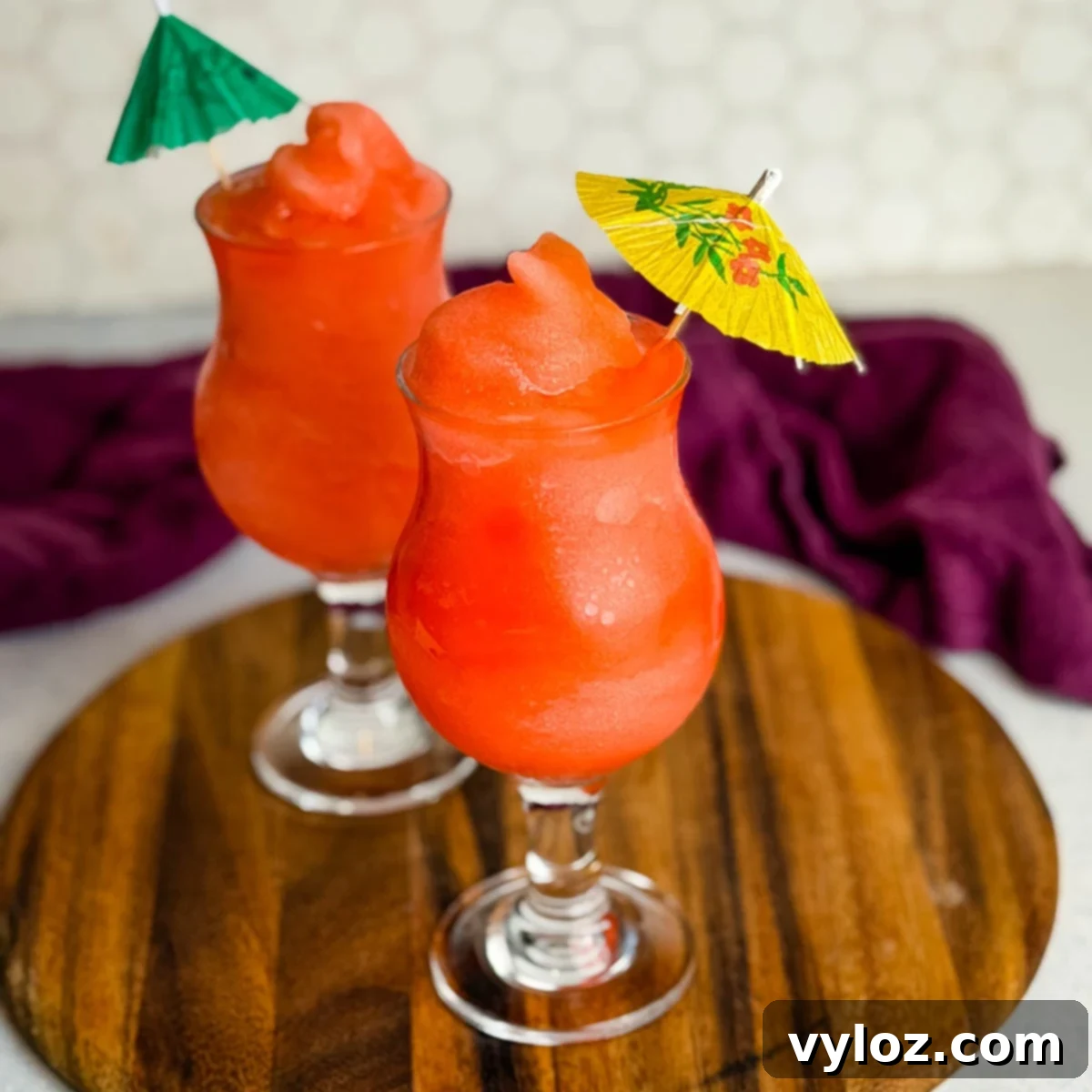 Two glasses of frozen Ninja Slushi Rum Punch topped with mini paper umbrellas on a wooden board.