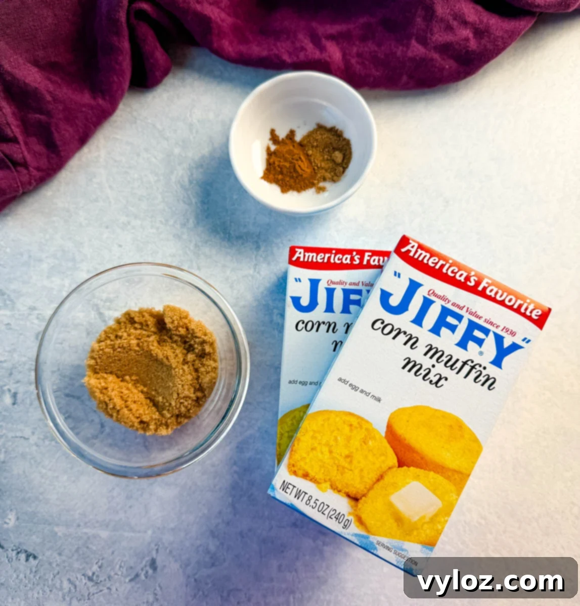 Ingredients for Jiffy sweet potato cornbread muffins including boxes of Jiffy Corn Muffin Mix, brown sugar, cinnamon, and nutmeg arranged on a light countertop with a purple kitchen towel.