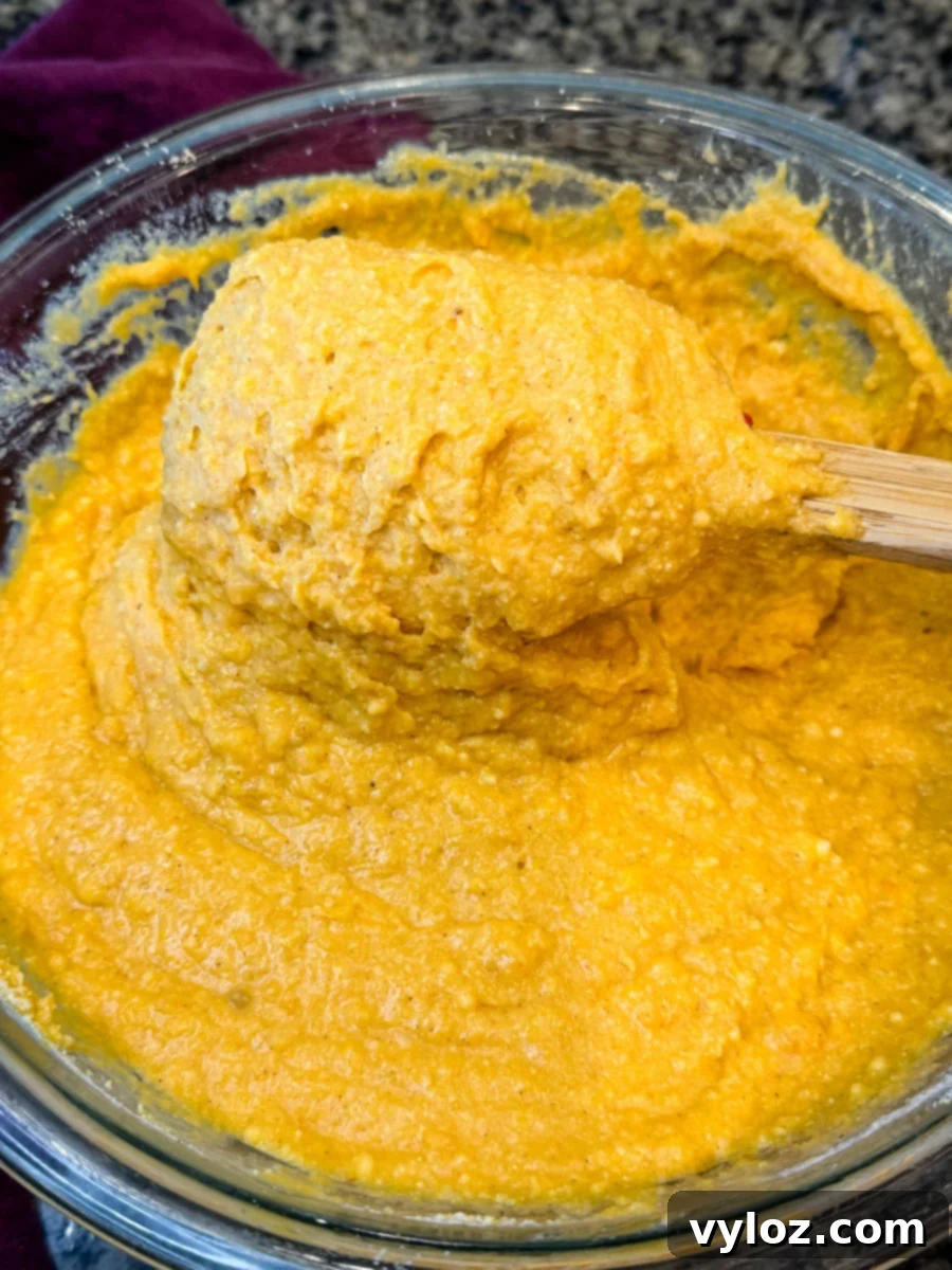 A bowl of thick, smooth sweet potato cornbread batter being stirred with a wooden spatula, showing its rich golden-orange color and creamy texture.