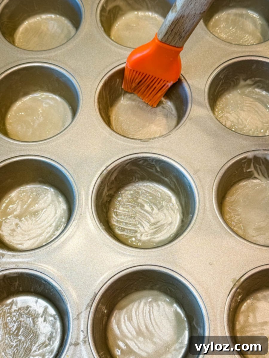 A muffin tin being greased with melted butter using an orange silicone brush, preparing the pan for baking cornbread muffins.