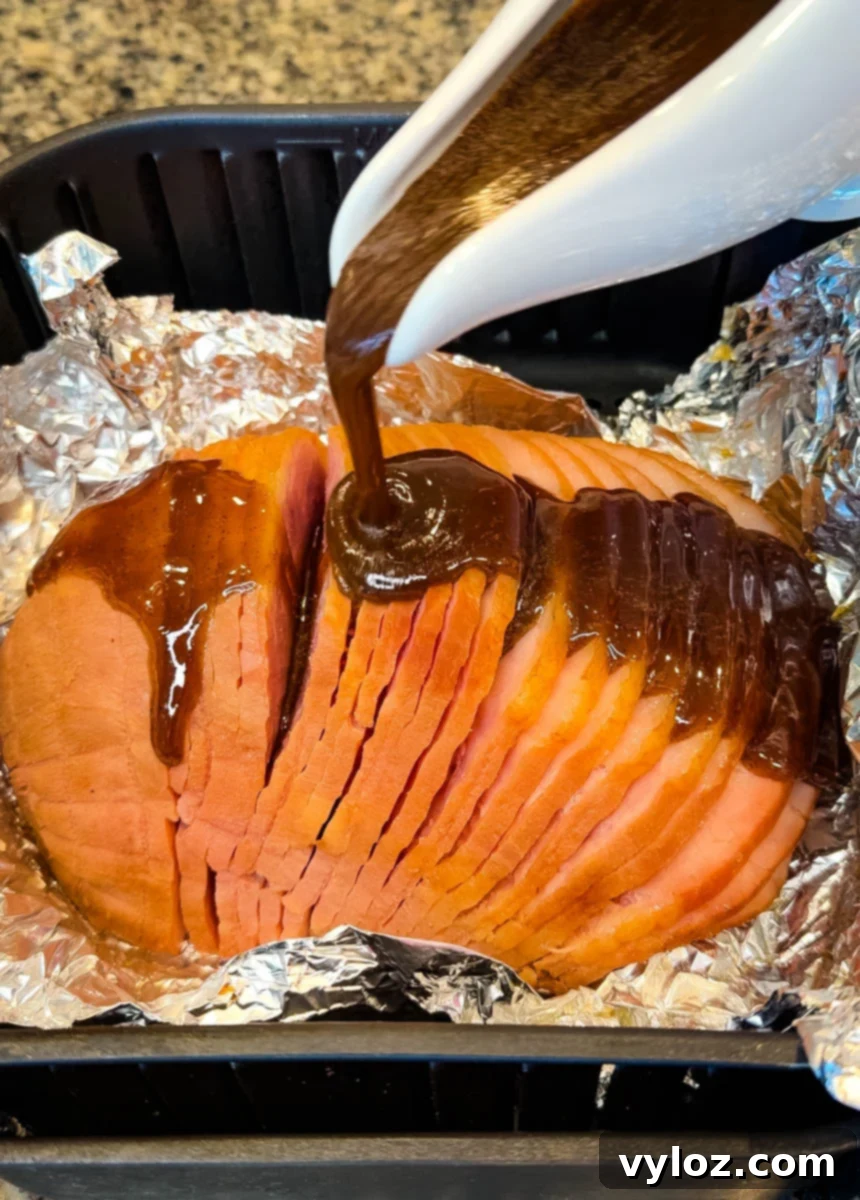 Honey-Kissed Air Fryer Ham 7 Thick brown glaze being poured over a sliced ham sitting on foil inside an air fryer basket, coating the ham for caramelization.