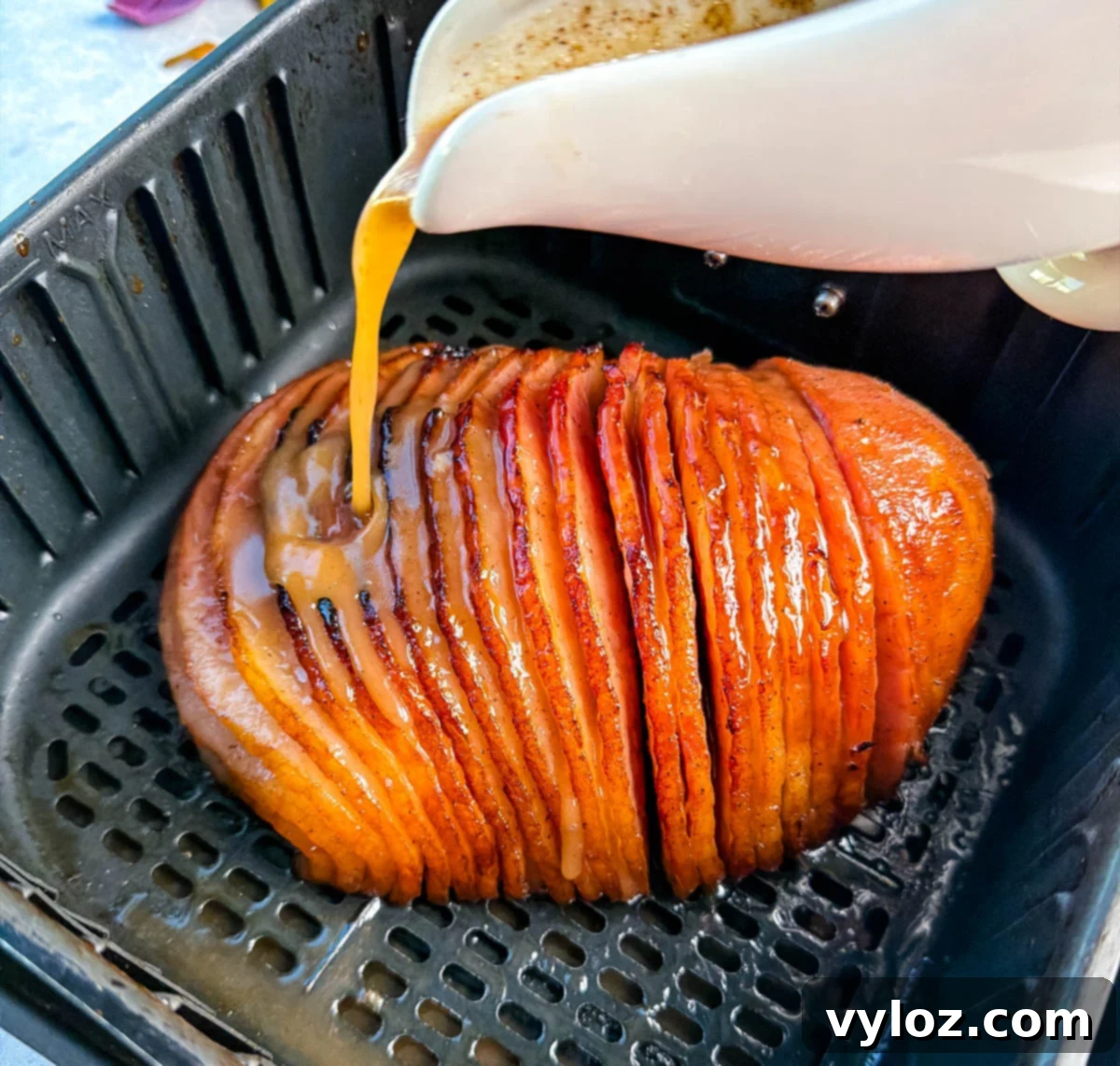 Honey-Kissed Air Fryer Ham 9 Honey glaze being poured from a white pitcher over a spiral ham inside the air fryer basket, coating it evenly for a glossy finish.