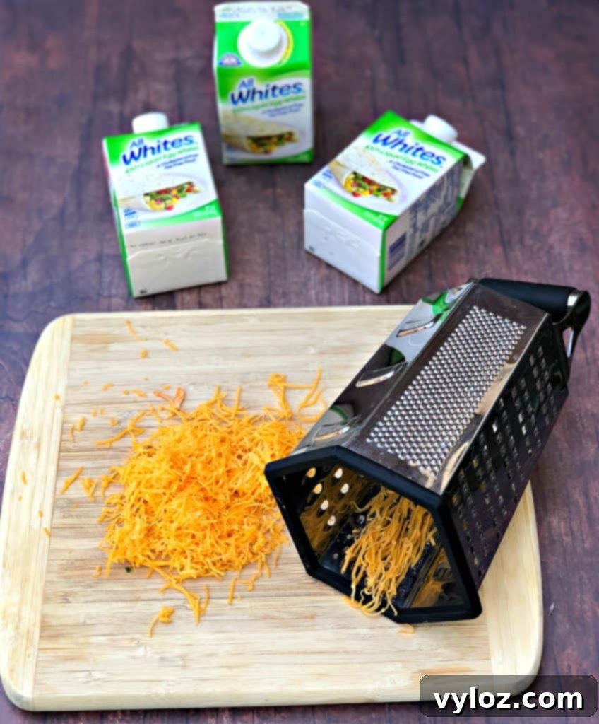 Keto Veggie Egg White Bites 3 A mound of freshly shredded cheddar cheese on a rustic wooden cutting board, with a grater resting nearby, highlighting a key ingredient for egg white bites.