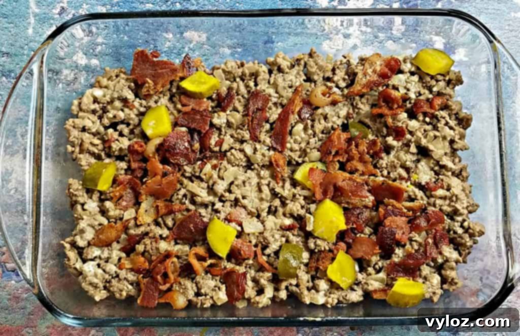 Keto Cheeseburger Bake 7 Ground beef mixture, topped with chopped pickles and crumbled bacon, ready for the creamy liquid layer in a baking dish.